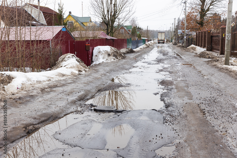 Obraz premium A muddy, puddled rural street lined with fences, bare trees and snow piles