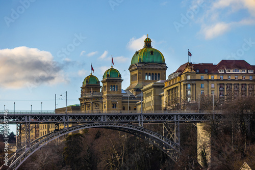 The grand domes of the Swiss Federal Palace rise majestically above the intricate steel arches of the Kirchenfeldbrücke, reflecting in the calm waters of the Aare river under a crisp blue sky.
