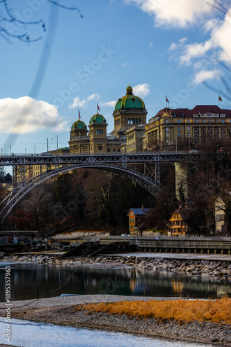 The grand domes of the Swiss Federal Palace rise majestically above the intricate steel arches of the Kirchenfeldbrücke, reflecting in the calm waters of the Aare river under a crisp blue sky.