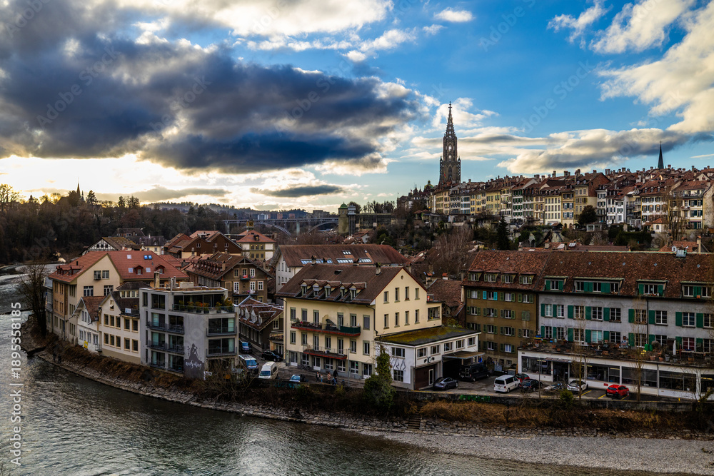 Obraz premium The historic houses of the Matte district line the banks of the turquoise Aare river, while the majestic spire of the Bern Minster rises above the city’s rooftops under a dramatic, cloud-filled aftern