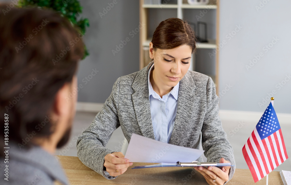 © Studio Romantic - Consular visa interview and immigrants documents discussion of female lawyer and man at office desk with US flag. USA immigration application and paperwork review at meeting by embassy employee