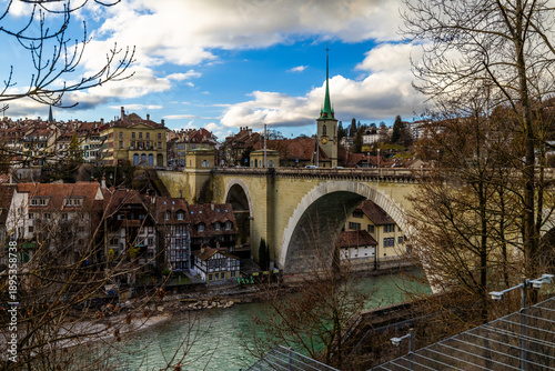 The impressive stone arches of the Nydeggbrücke span across the turquoise Aare river, connecting the historic Nydegg Church and the charming timber-framed houses of Bern's lower Old Town under a dynam