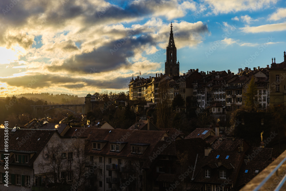 Obraz premium The dramatic silhouette of the Bern Minster dominates the skyline as golden evening light breaks through heavy clouds, illuminating the rooftops of the old city and the distant arched bridge.