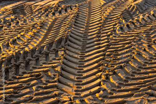 Close-up texture of heavy tire tracks on a sandy beach in sunset light. Pattern of protector imprints on the shore during low season coastal maintenance. Abstract industrial background in Turkey.