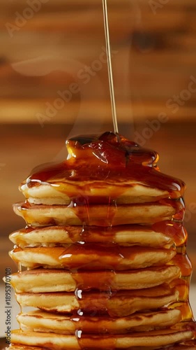 A tall stack of golden pancakes receives a generous pour of glistening syrup