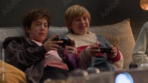 Medium shot of two Caucasian boys resting on sofa, chatting and joking while playing co-op videogame