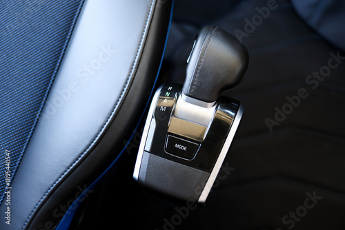 
Close-up of a modern automatic car gear shift lever with leather handle and chrome details, showing drive mode selector inside a stylish vehicle interior with dark upholstery and blue accents.