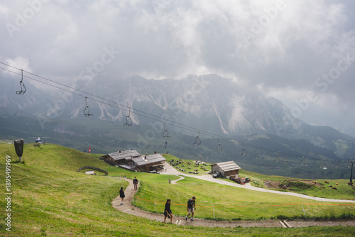 Εκτύπωση καμβά Seceda, Dolomites, Italy — alpine meadow with footpath and hikers, empty chairlift line cutting across the scene, mountain huts below cloud-covered peaks; summer hiking and tourism landscape