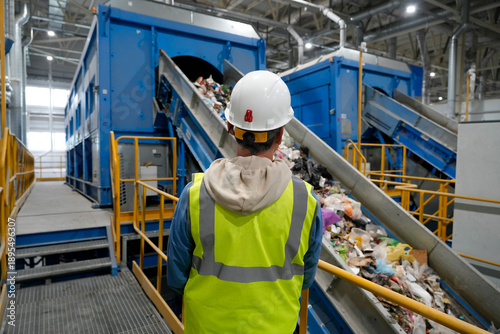 Obraz na plátně Male worker in a reflective vest and white helmet against the background of waste processing and sorting equipment on a conveyor belt, monitoring the quality of work of employees and personnel