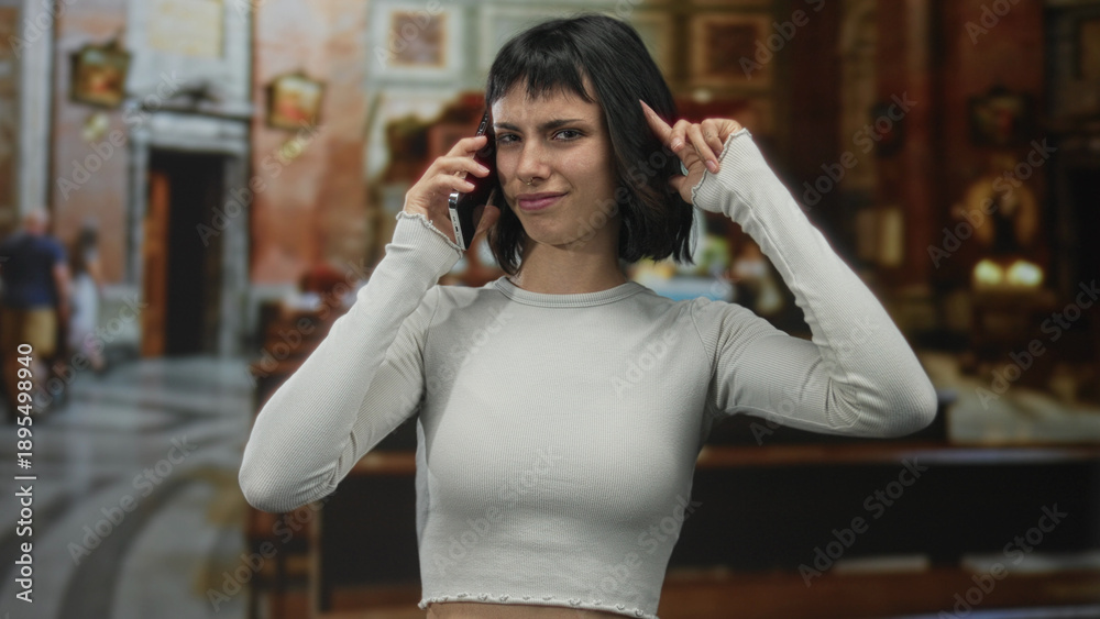 Fototapeta premium Hispanic woman holds smartphone while finger pointing to head gesture in building interior; frustration.