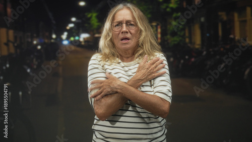 Fotografía Senior woman with folded arms and pursed lips standing under streetlights on a quiet urban street at night with hunched shoulders and bowed head; vulnerability solitude
