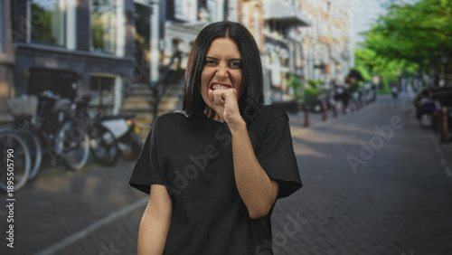 Bild auf Leinwand Woman biting her hand knuckle on a cobblestone street in front of bicycles and buildings, mouth clenched in a nervous gesture; anxiety uncertainty