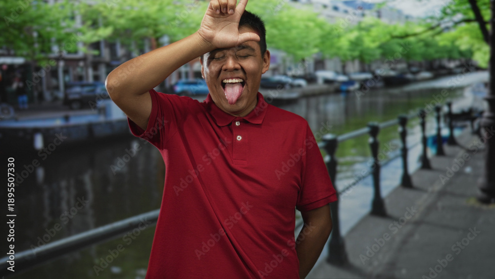 Fototapeta premium Man wearing red polo sticks out tongue and forms l with hand to forehead on street by canal in amsterdam; playful mischief.