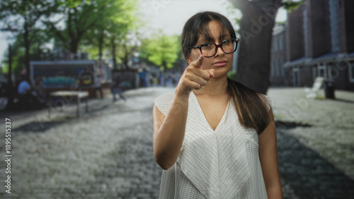 Woman in white blouse points finger at camera on cobblestone street by brick building; accusation challenge.