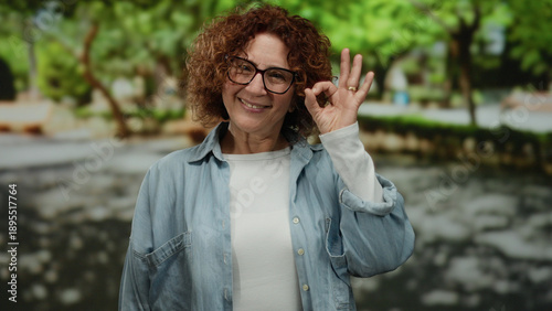Woman smiling in a park, wearing glasses and casual clothes, gesturing thumbs up with a bright expression, surrounded by green trees on a sunny day.