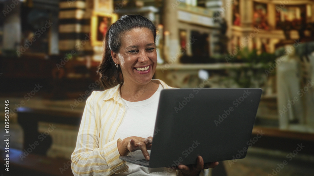 Fototapeta premium Woman holding laptop and tapping the keyboard with her right hand in an ornate church interior, smiling while looking at the screen; joy.