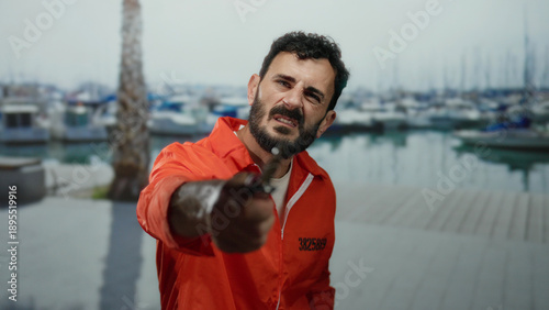 Картината върху платно Hispanic man with beard in prison jumpsuit holding knife at seaside promenade with boats and palm tree visible