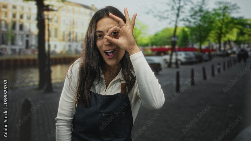 Fototapeta premium Woman with exposed face wearing denim apron and white shirt, winking and making ok sign to eye on amsterdam street by canal; playful confidence.