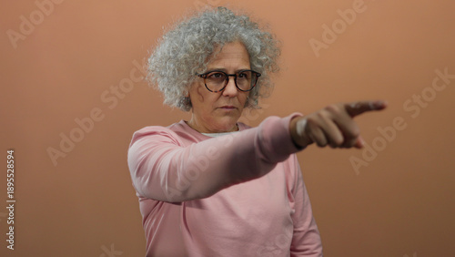 Photos Middle-aged woman with glasses and curly grey hair pointing with focus, wearing a pink sweater against a warm brown background, isolated indoors