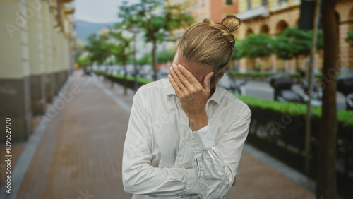 Tableau sur toile Blond man with long hair wearing a white shirt and hand covering face while standing on a sunlit street; frustration
