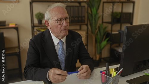 Obraz na plátne Senior businessman in suit and glasses contemplating at desk in modern office interior, holding a pen, surrounded by plants and office supplies, representing leadership and thoughtfulness