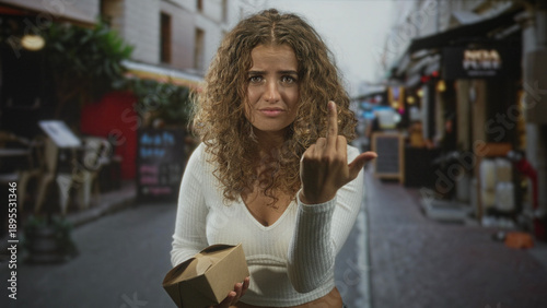 Bilde på lerret Young woman holding a takeout box shows middle finger with hand gesture on a busy cafe street; defiance