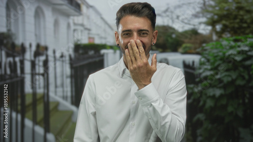 Obraz na plátně Young hispanic man covering mouth with hand on urban street by white townhouse facade; lighthearted joy