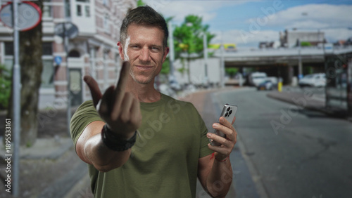 Man in green tshirt shows middle finger while holding a smartphone on a city street near a building and parked car; defiance confrontation.