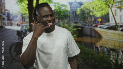 Man making call gesture with hand and smiling on street by a canal with bicycles and cars; friendly invitation.