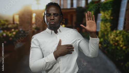 Tableau sur toile Young african american man with glasses wearing white shirt with hand on chest and raised palm on cobblestone street by brick building and planters; honor pledge respect