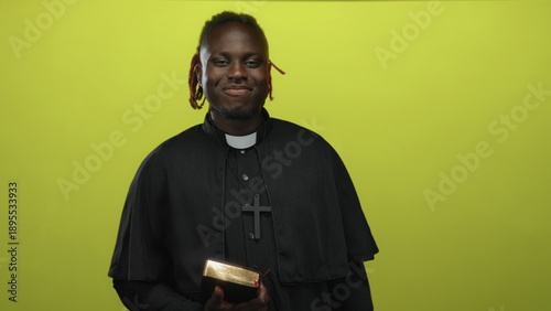 Fotografía Man priest holding a bible in studio against lime green wall, smiling and presenting the book while wearing clerical collar; faith hope serenity