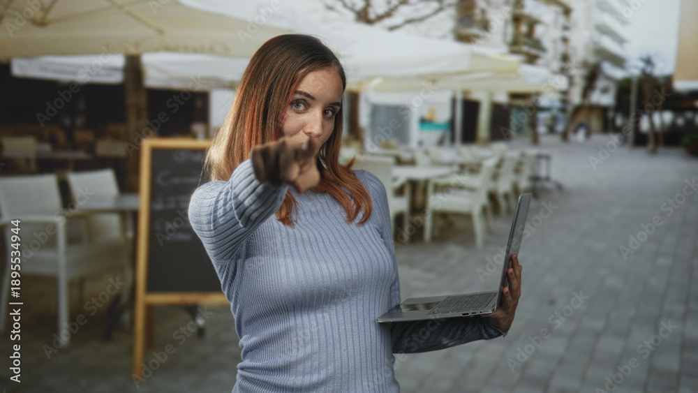 Fototapeta premium Woman pointing finger toward camera while holding laptop on a street cafe terrace beside a chalkboard menu and patio umbrellas; confidence.