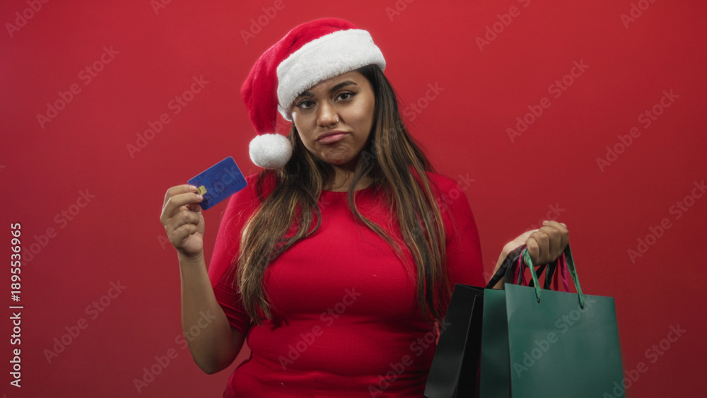 Obraz premium Woman holding shopping bags and a creditcard, wearing a santa hat and frowning while holding up the card, studio; holiday frustration.