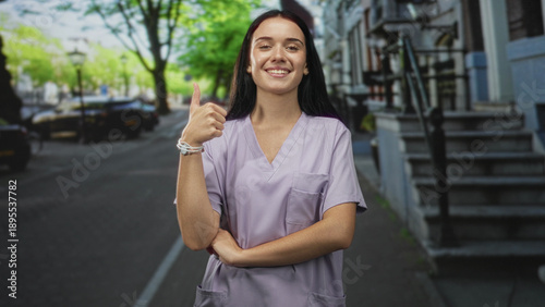 Young nurse in lilac scrubs gives thumbs up with visible hand and arm on street beside building stoop; confidence care reassurance. Free Download Young nurse in lilac scrubs gives thumbs up with visible hand and arm on street beside building stoop; confidence care reassurance. - GFXLoad