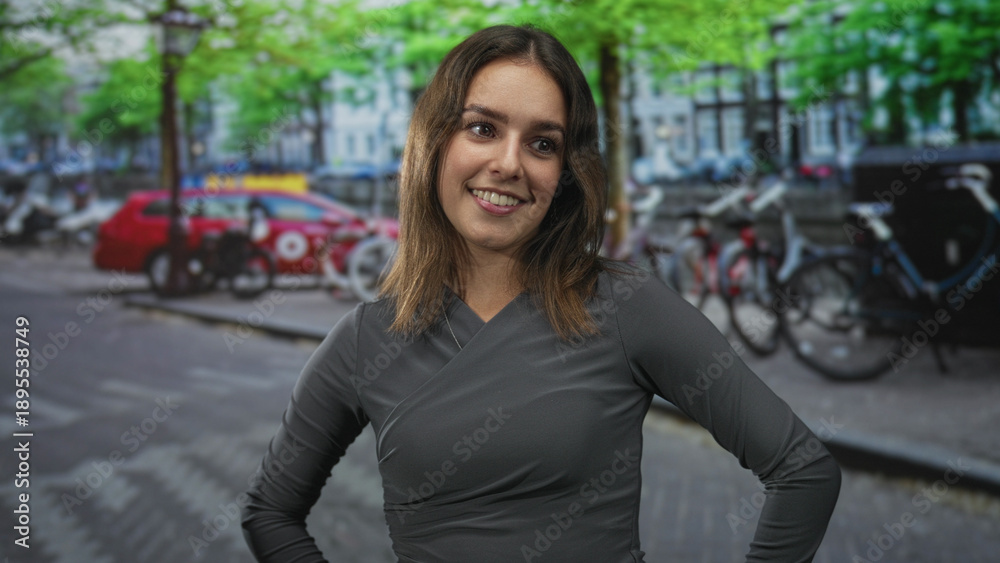 Fototapeta premium Woman smiling with hands on hips on a city street with parked bikes, a red car and trees; confidence warmth curiosity.
