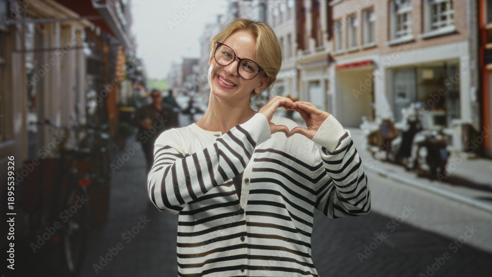 Fototapeta premium Woman with blond hair and glasses forming a heart gesture with hands while smiling in a striped sweater on a cobbled street lined with shops and bicycles; affection connection.