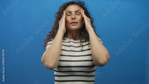 Foto Young hispanic woman in a striped shirt gently touches temples and frowns with closed eyes in a blue studio; stress