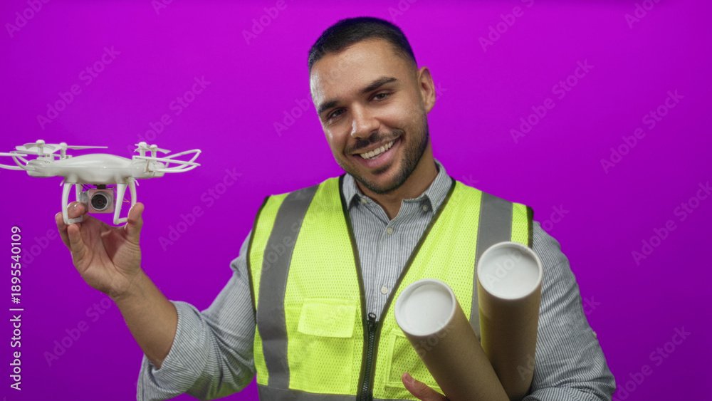 Fototapeta premium Young man holds drone in purple studio with rolled blueprint tubes; determination inspiration progress.