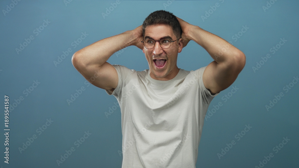 Naklejka premium Man holding hands behind head, mouth open and wearing glasses in a studio with a blue backdrop and light gray t shirt; surprise reaction.