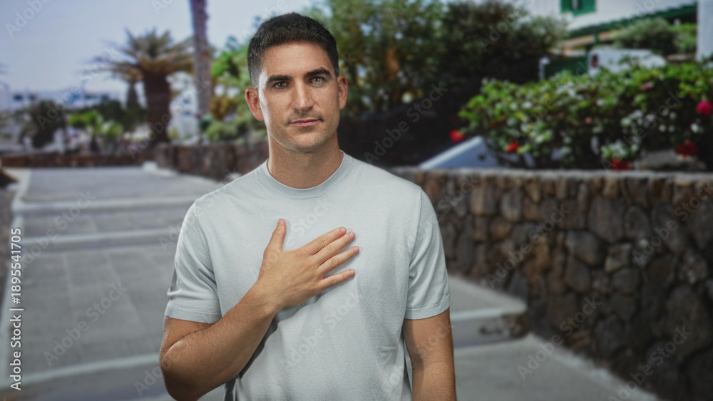 Fototapeta premium Man with hand on chest on street by stone wall and steps in city sunlight, outdoors and standing near plants; sincerity reflection.