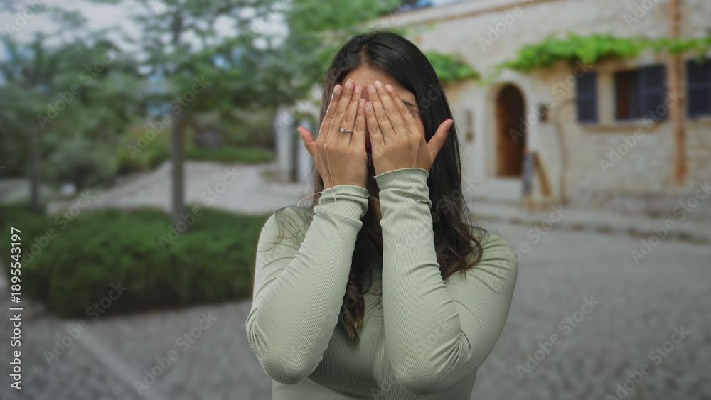 Fototapeta premium Young hispanic woman in olive top covers face with hands smiling in front of stone building; playfulness.
