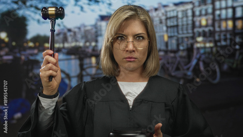 Canvastavla Young blonde judge wearing black robe raises gavel on canal street in amsterdam with focused gaze; justice authority