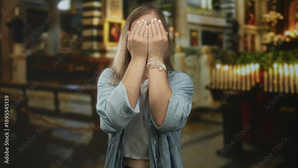 Fototapeta premium Woman blonde covers eyes with hands and makes playful faces while standing in a church building interior near candles and pews; playful surprise.