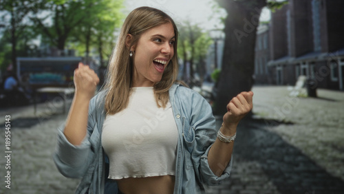 Foto Young blonde woman wearing white crop top clenching fists on cobblestone street; triumph victory celebration joy