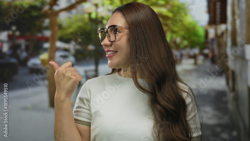 Young woman wearing eyeglasses and casual white top shows thumbs up gesture on sunny urban street lined with trees and blurred traffic; optimism.