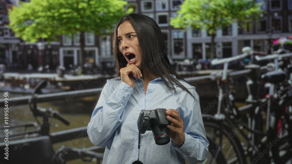 Fototapeta premium Woman holding camera and pulling at shirt while grimacing on street in amsterdam canal area; discomfort travel fatigue.