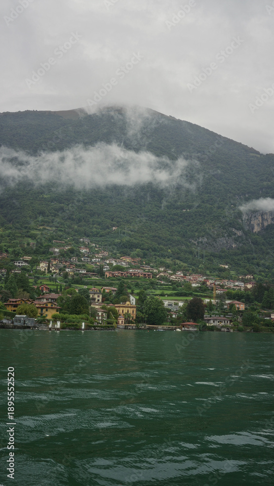 Obraz premium Lakeside village with waterfront buildings, rippling emerald lake water and low clouds clinging to the mountain slopes; serenity.