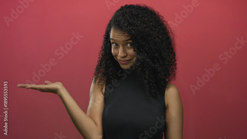 Tableau sur toile Young hispanic woman extends hand palm up and points finger at camera in red studio; warm invitation