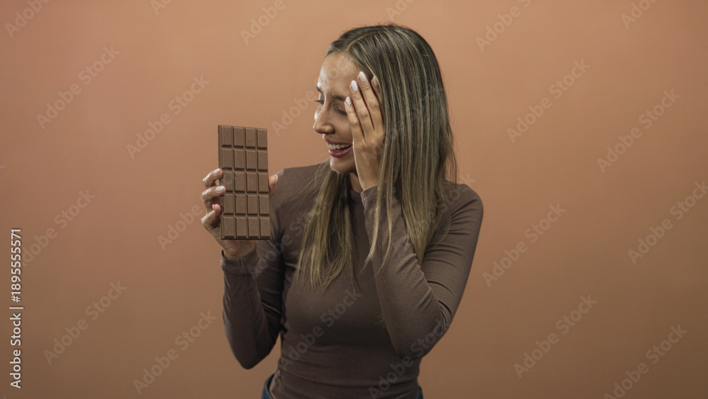 Φωτογραφία Young hispanic blonde woman smiling while holding a large chocolate bar and covering one eye with her hand in a peach studio; playful indulgence