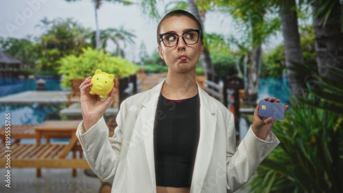 Εκτύπωση καμβά Woman holding a yellow piggy bank and credit card, puckers lips while weighing options beside a pool building; financial choice thoughtful